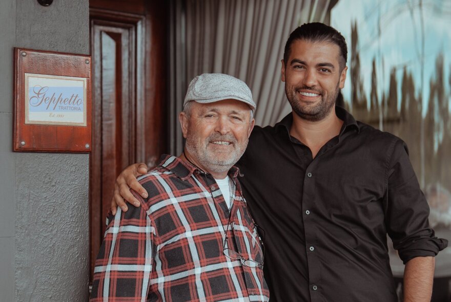 Two men standing and smiling, the taller, younger man with his arm around the older man wearing a cap.
