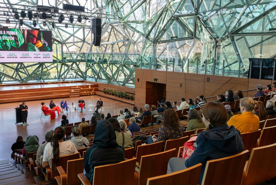 A group of people are watching a talk in a large glass auditorium. 