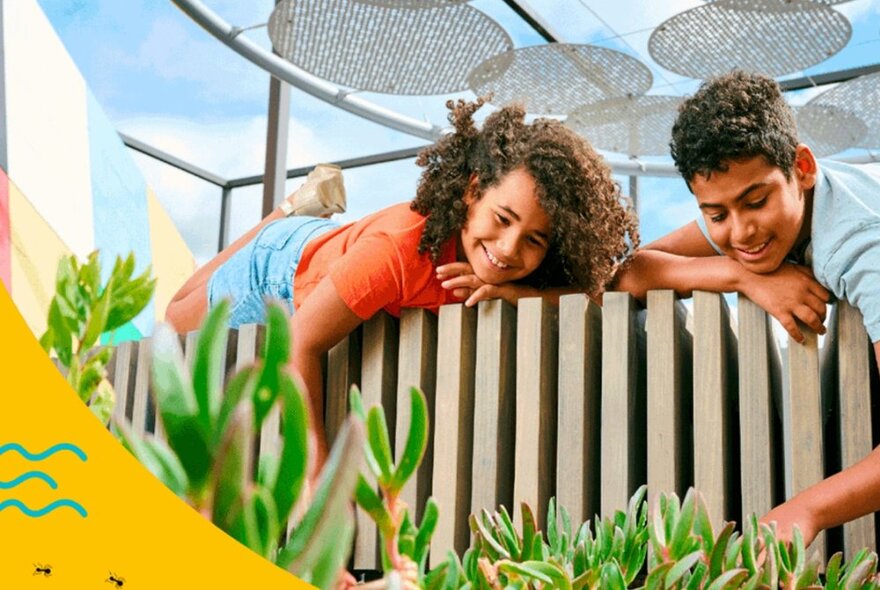 Two kids leaning over a wooden picket fence, touching the green plants below, blue sky and circular sunshades above them.