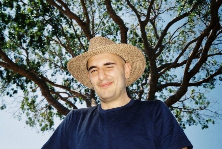 Stacy Gougoulis, a man in a dark t-shirt and large sunhat, photographed from below, with a tree and the sky visible behind him.