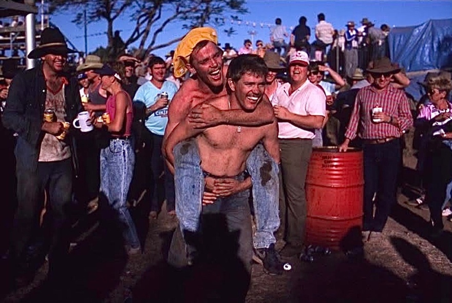 A crowded scene from an outdoor event with a shirtless man giving another a piggyback. 