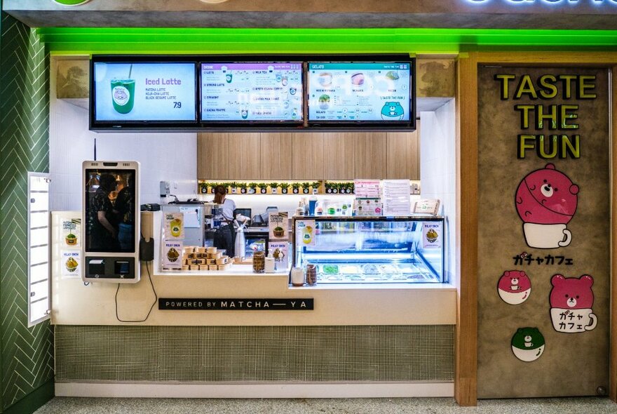 A takeaway food counter with display screens on the back wall, a refrigerated glass display and cute signage of Japanese cartoon style characters and a logo that says 'TASTE THE FUN', written on a wall.