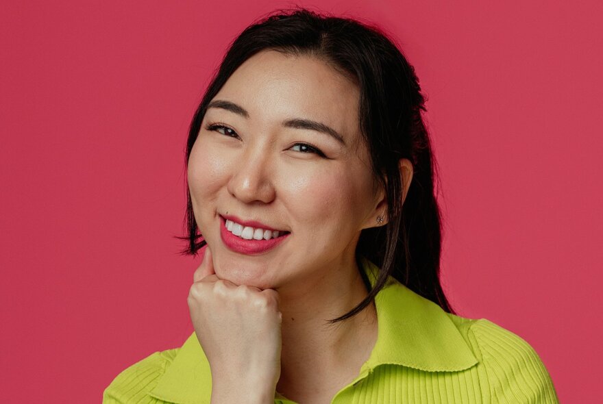 A smiling young woman with black hair tied back in a ponytail and her chin resting on her knuckles.