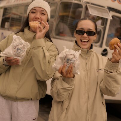 Two friends wearing light coloured jackets enjoying hot donuts near the donut van; they each hold a paper bag in one hand and a donut in their other hand.