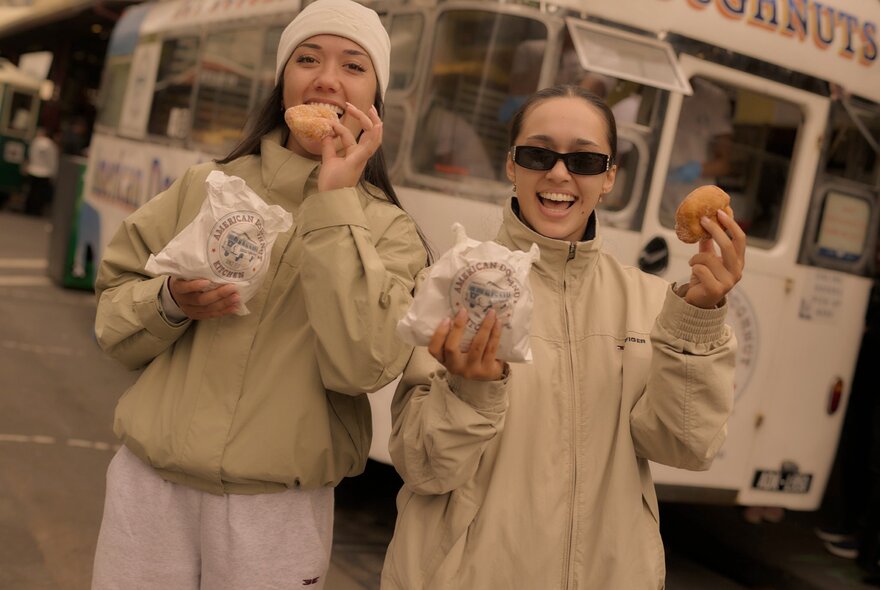 Two friends wearing light coloured jackets enjoying hot donuts near the donut van; they each hold a paper bag in one hand and a donut in their other hand.