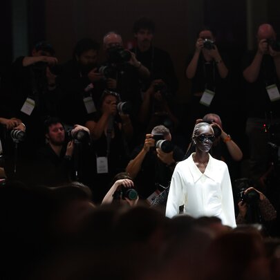 A runway model wearing a tailored white long-sleeved shirt walks toward a darkened audience filled with photographers taking photographs.