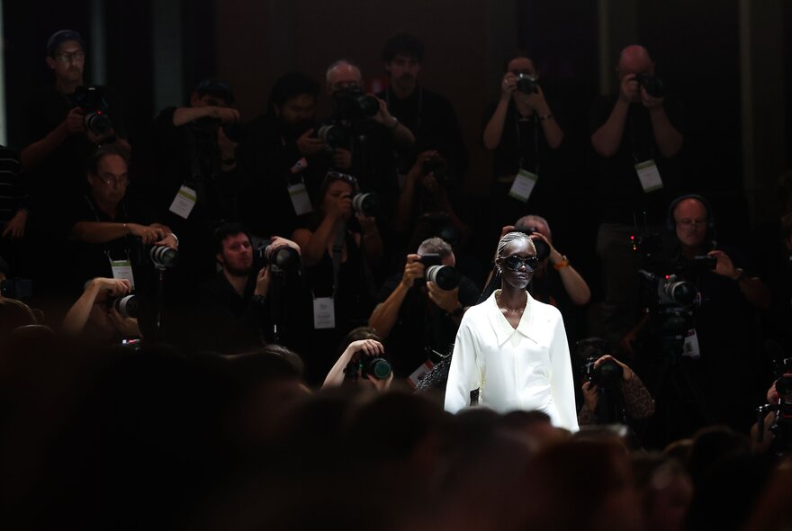 A runway model wearing a tailored white long-sleeved shirt walks toward a darkened audience filled with photographers taking photographs.