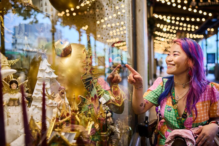 A smiling woman pointing at a shopfront window, garlands of fairy lights line the street behind her. 