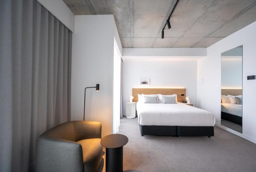 A spacious room at the Nesuto Docklands Apartment Hotel, with a king size bed, small sofa, lamp and grey curtains.