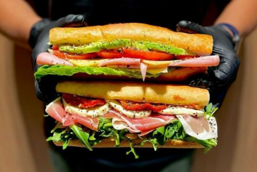 A pair of delicious looking filled french baguettes being held one above the other by two hands in black gloves.