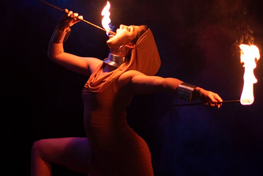 A woman in a hooded red costume holding a flaming fire-eating apparatus against her tongue, with a second baton in her other hand.