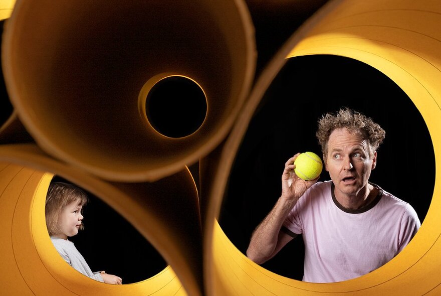 An adult and a child in an abstract yellow sculptural space, the adult holding a ball to his ear.