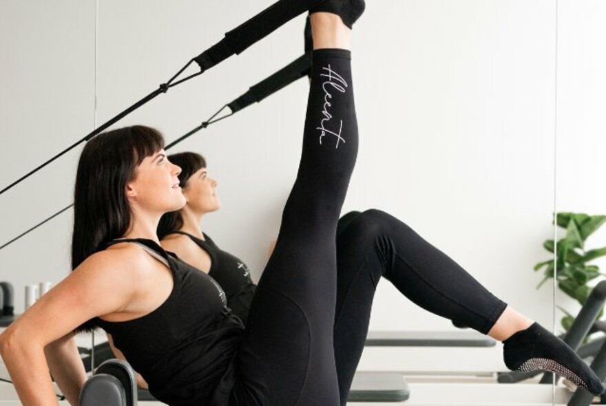 Woman in black active wear on a Pilates Reformer machine, with leg raised and pushing against an elastic strap. 