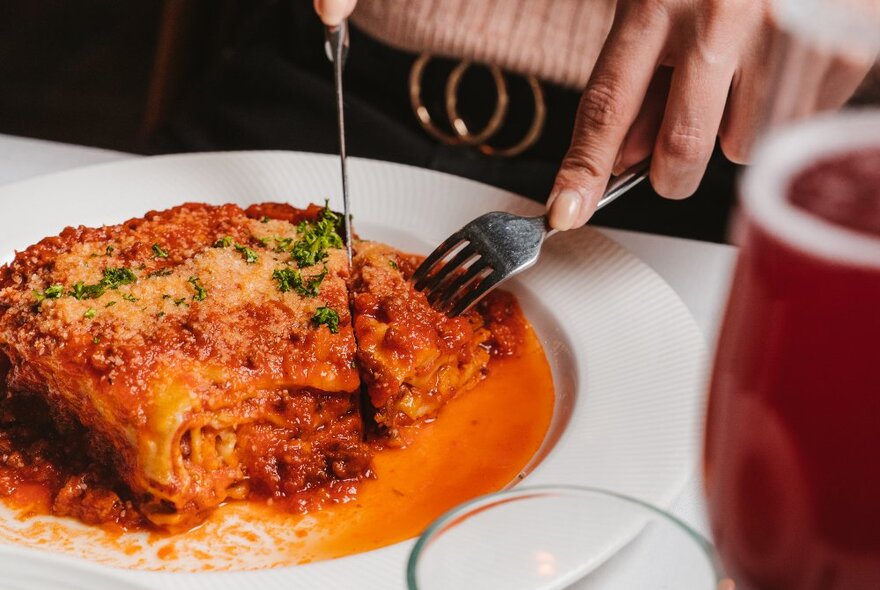 Hands holding cutlery cutting into a serve of lasagne with tomato sauce in a white bowl.