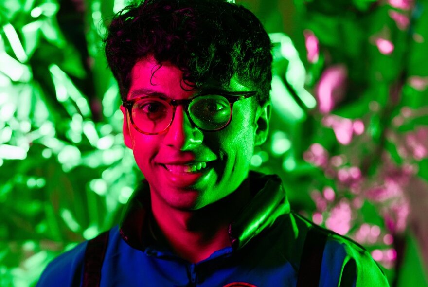 Close-up of face of a smiling man wearing glasses in half shadows, in front of lush greenery.