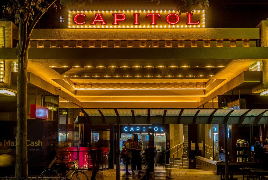 Outside an art deco movie theatre at night with a glowing red and yellow sign saying CAPITOL. 