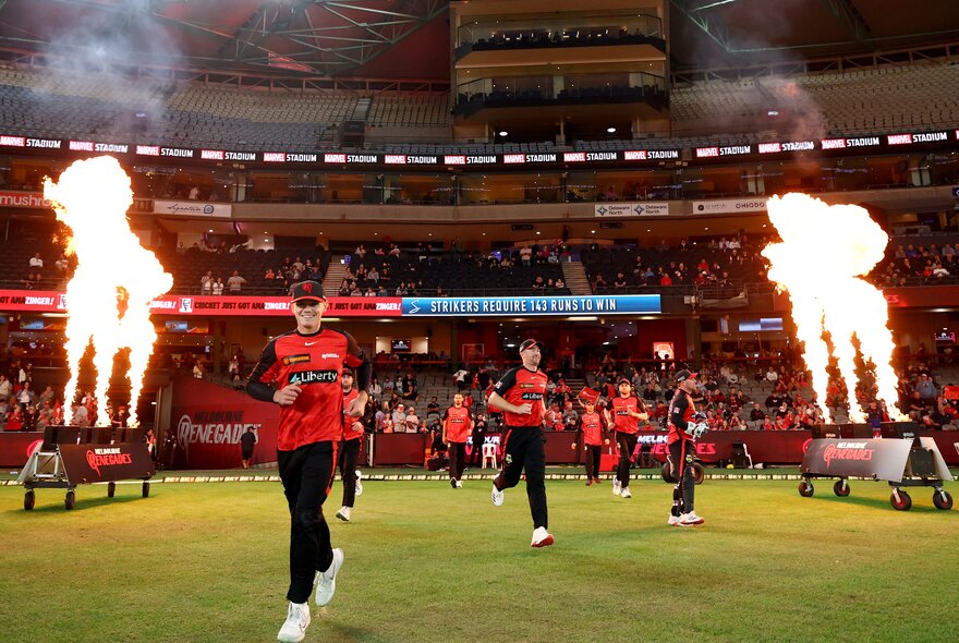 Melbourne Renegades BBL players walking out on the cricket field, past jets of flames, with cricket fans in the stadium seats in the background.