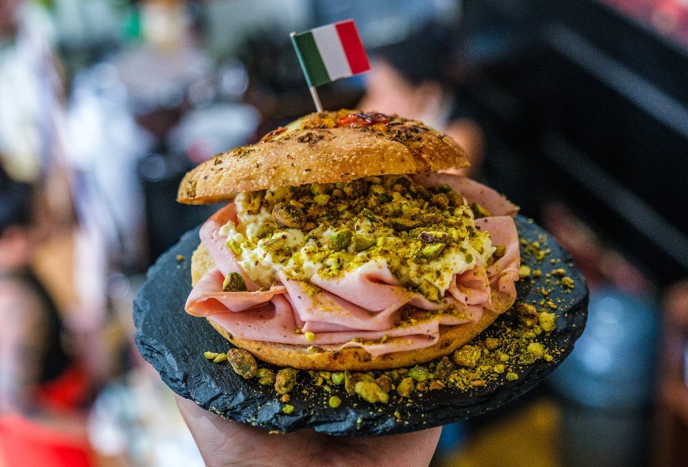 A focaccia sandwich filled with mortadella, cheese and pistachios, and topped with a small Italian flag.