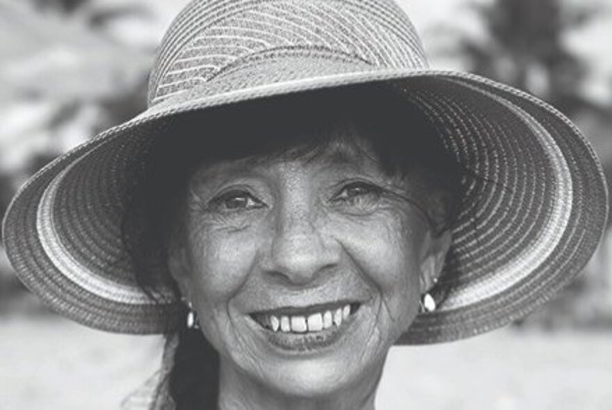 An older woman smiling, wearing a large straw sunhat, her hair loosely pulled back off her face; black and white.