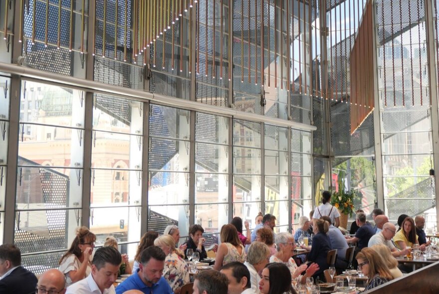 The interior of Taxi Kitchen at Transport Hotel, with tables of patrons eating and drinking at long tables, surrounded by high floor to ceiling windows.