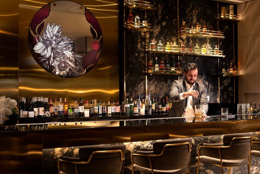 Man mixing cocktails behind an opulent bar, a large mirror and a wall of bottled drinks displayed behind the bar.