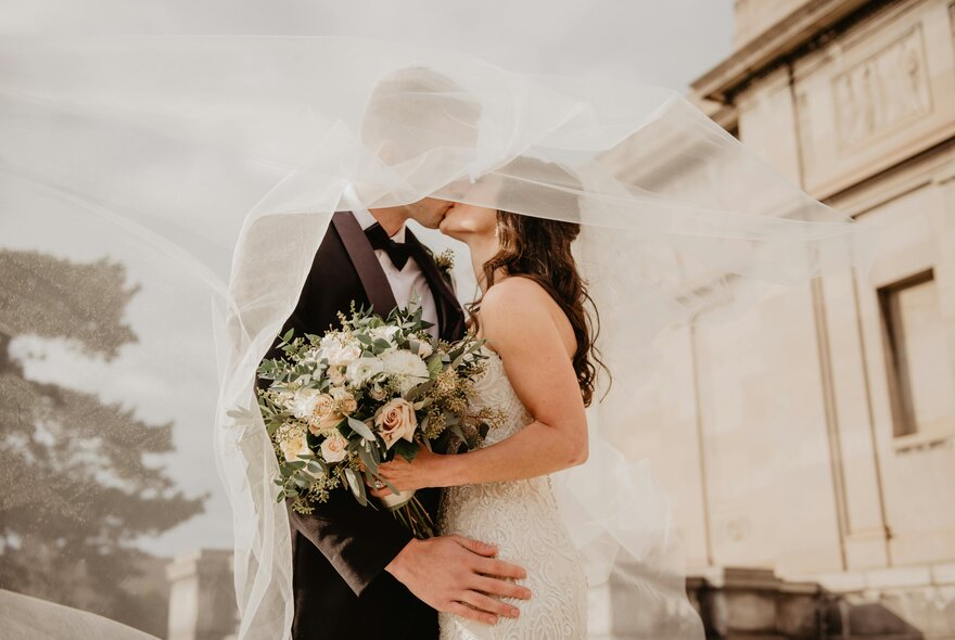 A bride and groom in an embrace kissing, wrapped in the bride's veil, with a stately building in the background.