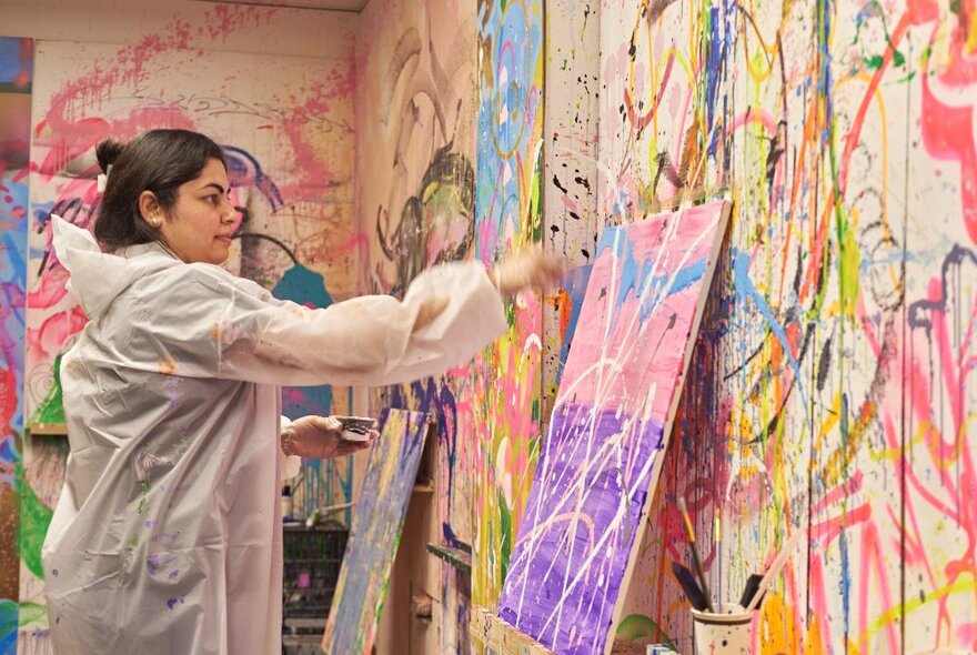 A  person in protective plastic clothing painting onto a canvas that rests on a ledge against a paint splattered wall.