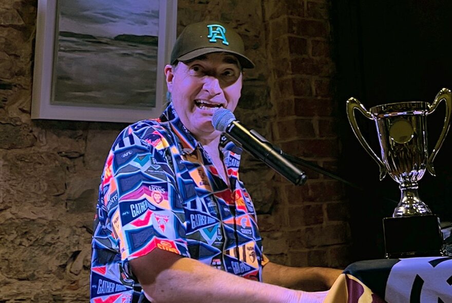 A man in a colourful shirt and baseball cap singing into a microphone, with a large gold trophy behind him.