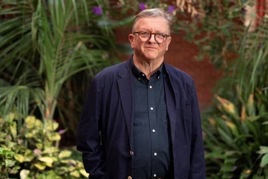 Academic Ross Jones, wearing spectacles, navy shirt and jacket, standing amongst plants in a walled garden.