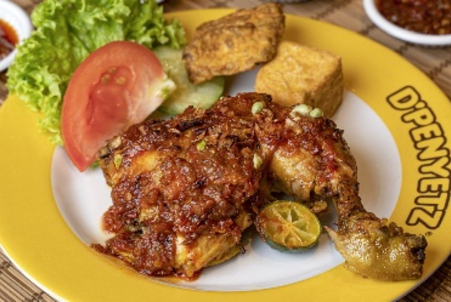 Fried chicken with tofu cubes and salad served on a plate that has the words D'Penyetz written on the yellow rim.