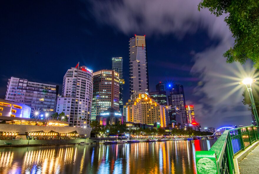 Vibrant view of the Melbourne's Southbank and the Yarra River at night with buildings, including the Langham Melbourne, illuminated and their colorful lights reflecting on the water's surface.