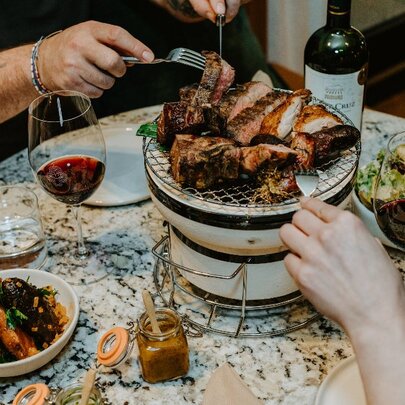 Two people enjoying an at-table Argentinian grill experience with chunky cuts of meat, sides and two glasses of red wine.