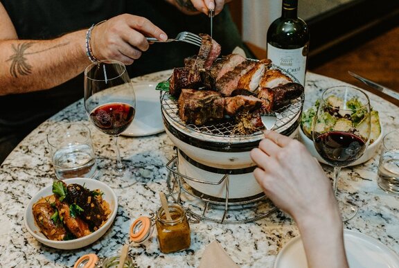 Two people enjoying an at-table Argentinian grill experience with chunky cuts of meat, sides and two glasses of red wine.