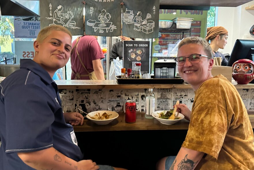 Two people look behind them and smile while sharing a meal on a small table.