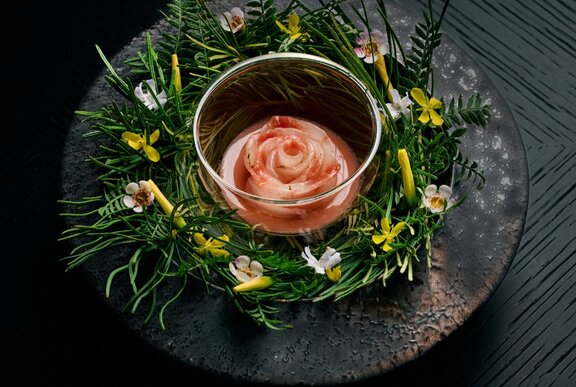 A swirl of kingfish sashimi inside a small glass dish surrounded by spring blossoms on a black earthenware dish.