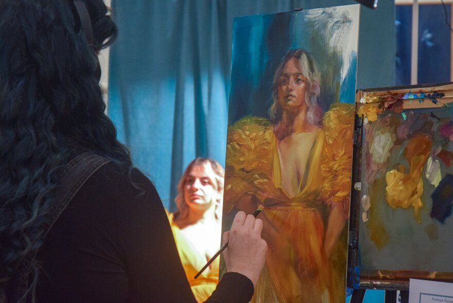 An artist painting the portrait of a woman seated in front of a bue curtain, wearing a yellow gown.