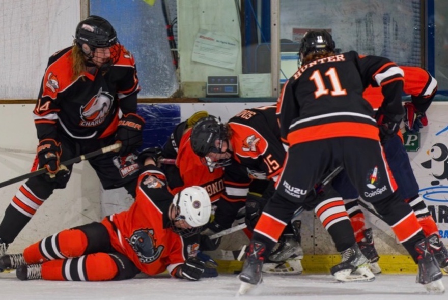 intense scramble for the puck along the boards during an ice hockey match involving players in black, orange, and red jerseys.