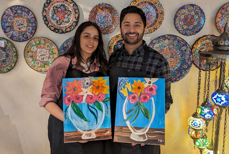 A man and a woman holding amateur acrylic paintings of vases with flowers, in a workshop with ceramic mosaic plates displayed behind.