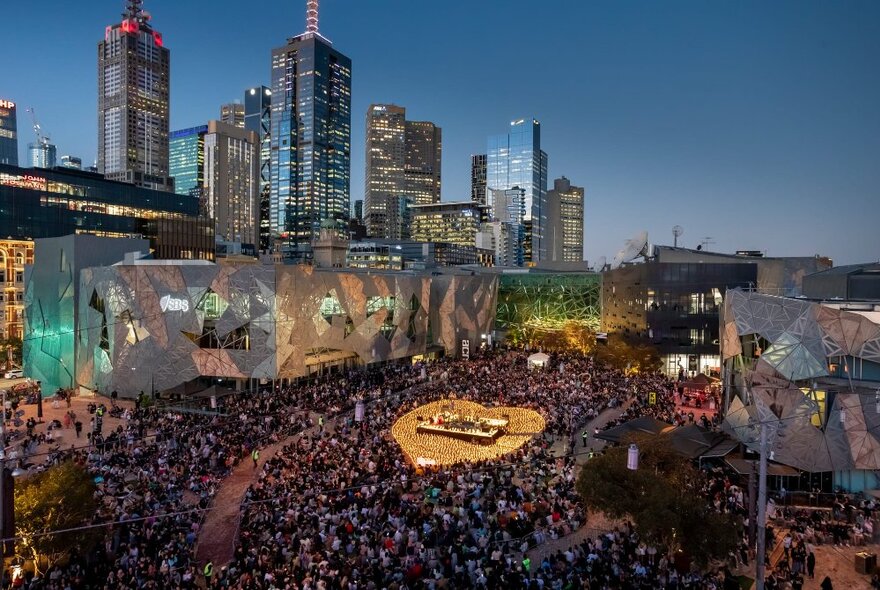 A drone shot of Fed Square at dusk, with a heart-shape of lights surrounding a central stage in the middle of a huge crowd.