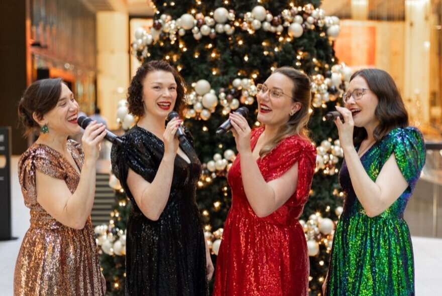 Singers in red, gold and green dresses, holding microphones in front of a decorated Christmas tree.