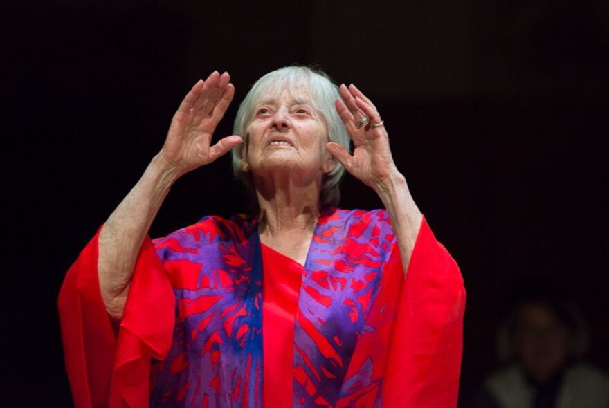 A grey-haired woman wearing a red and purple gown, her hands raised.