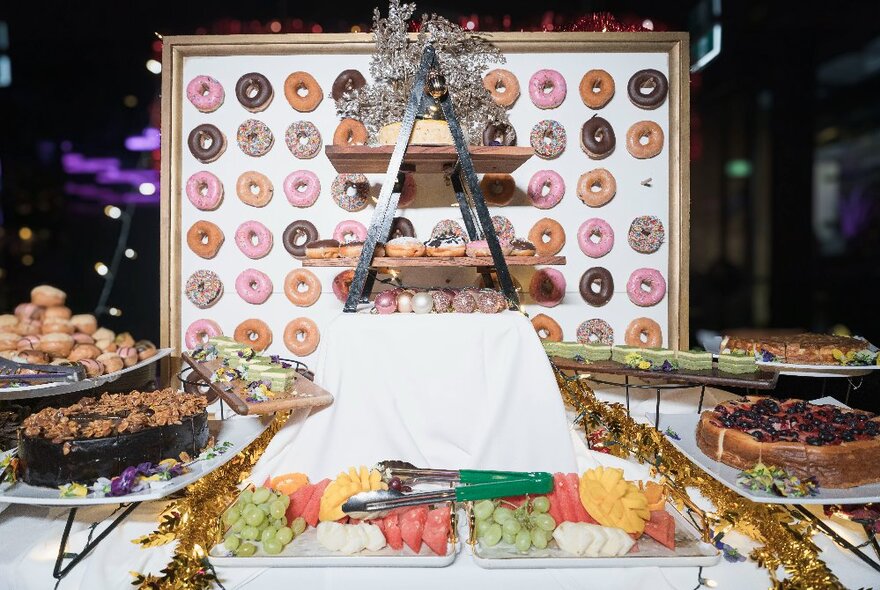 A decadent dessert buffet with a donut wall, tiered cake stand and fruit platters.