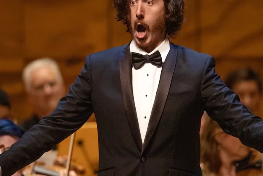 A male opera singer on stage in a tuxedo, singing with arms outstretched.