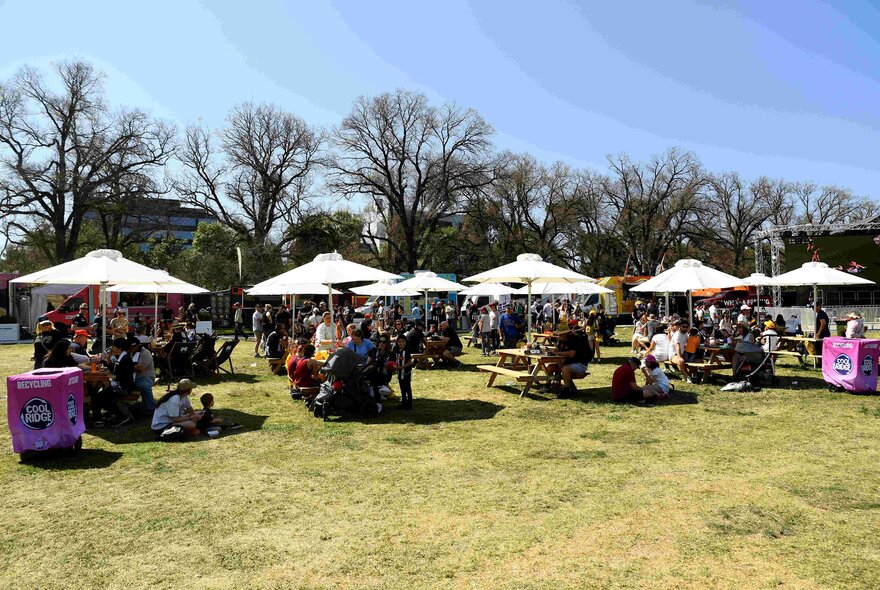 Grassy areas of Yarra Park set up with large picnic umbrellas and table, with people seated around them, as part of the AFL Footy Fest celebrations.
