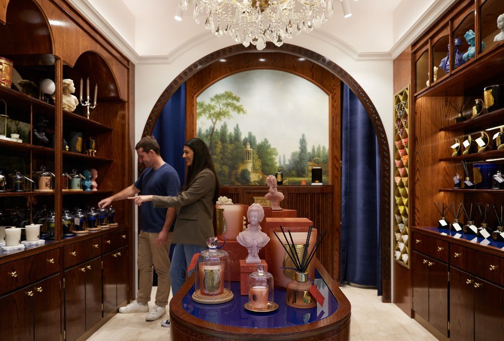 A man and a woman are pointing at a shelf of candles in a candle shop with antique wooden furnishings and dark blue accents.