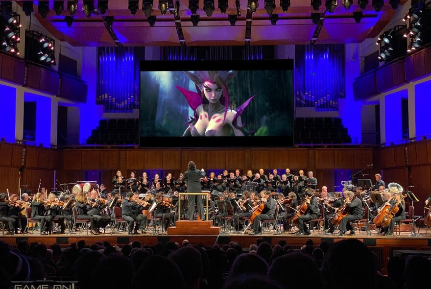 An orchestra performing live on stage while a animate game plays on the screen behind.