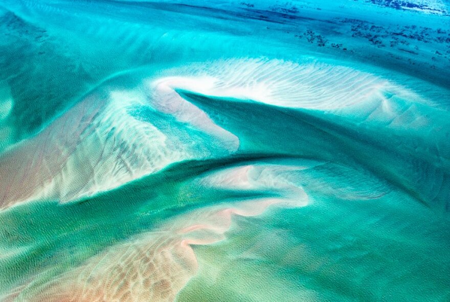 An aerial photograph over the ocean showing greens and blues and the curves of the same under the water. 