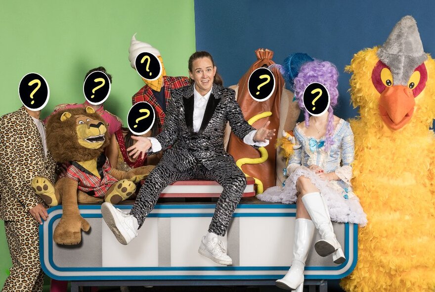Zoe Coombs-Marr on a bench with people in costumes such as a hot dog and bird, with question mark stickers over their faces. 