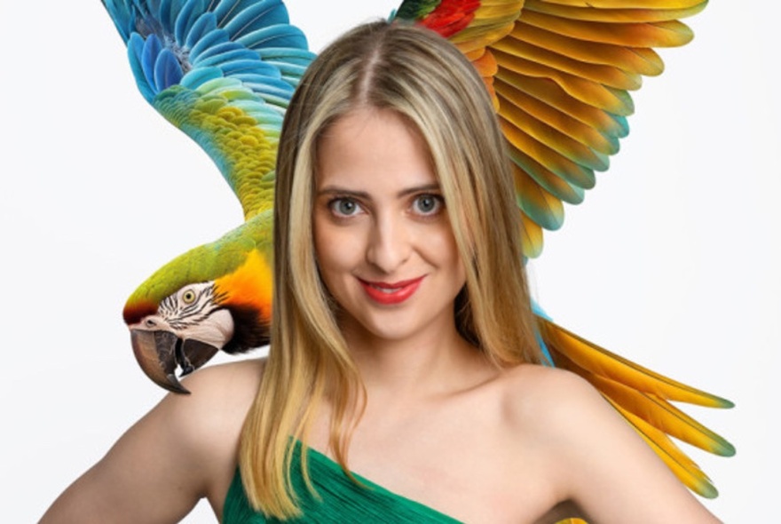 Smiling comedian Isabella Charlton, her straight long blond hair falling behind one bare shoulder and an image of a colourful toucan behind her head, as if resting on her shoulder.