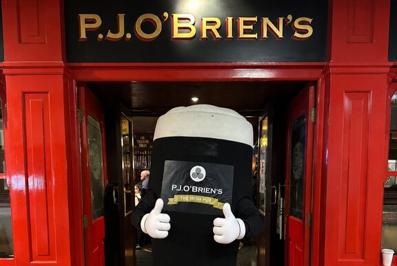 A Guinness pint mascot character stands at the entrance to P.J. O'Brien's Irish Pub framed by a vibrant red facade.